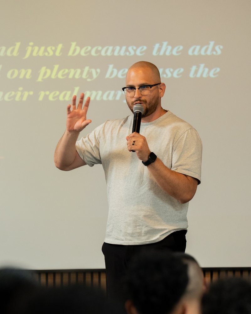 Emanuel Ruiz speaking into a handheld microphone at a live event, one hand raised mid-explanation, in front of a projected slide.